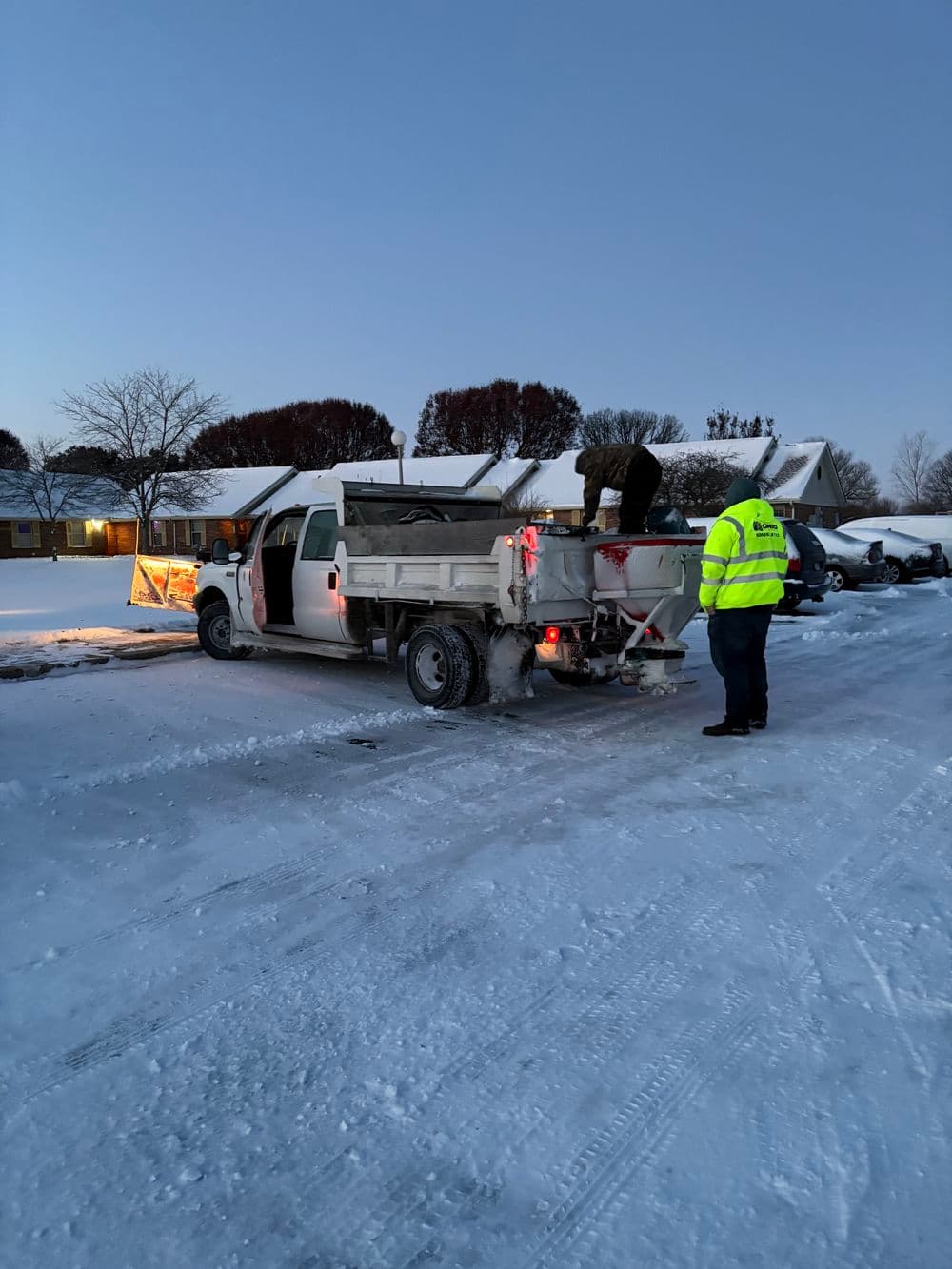 Snow plow truck unloading salt on a snowy road, with workers in safety vests nearby.