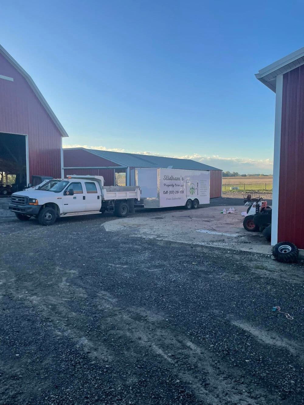 Delivery truck and trailer parked beside red barn in rural landscape during clear day.