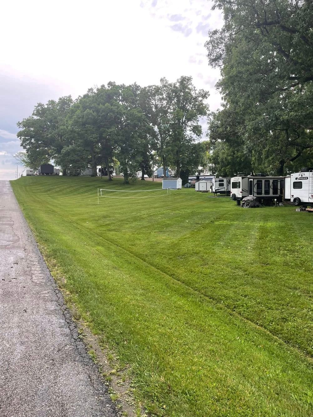 Grassy hillside with trees and parked RVs alongside a paved road under a cloudy sky.