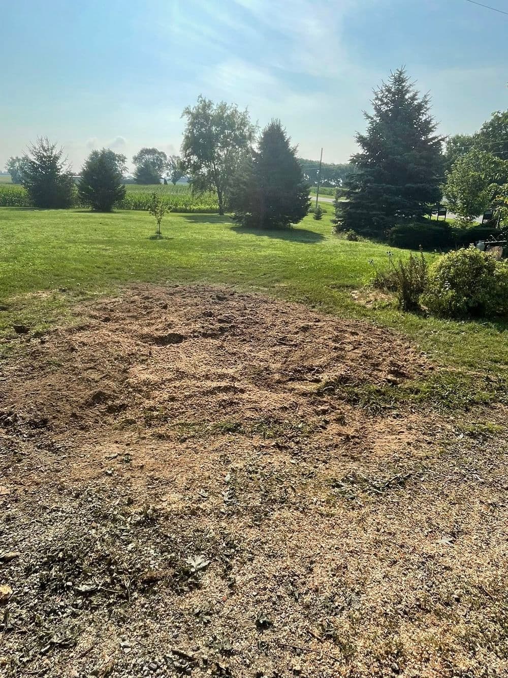 cleared land area for landscaping with trees and grassy field in background under blue sky