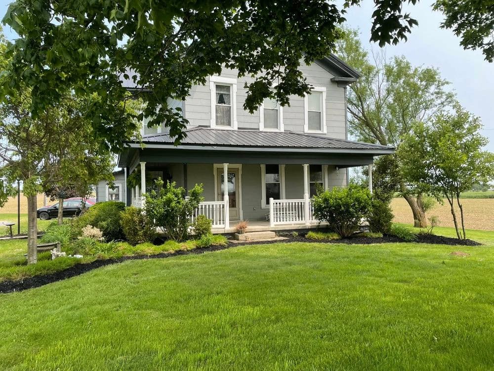 Two-story gray farmhouse with porch, surrounded by greenery and lush grass.